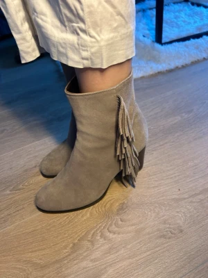 Beige mocka stövletter med fransar - Snygga beige stövletter i mocka med coola fransdetaljer på sidan och bred klack. De har en stilren siluett och spetsig tå som ger en trendig look. Perfekta för dig som vill ha något extra till outfiten.