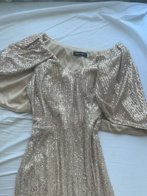 Glittrig champagnefärgad långklänning - Säljer en glittrig långklänning från Miss Ord i champagnefärg med paljetter över hela klänningen. Klänningen har v-ringning och vida, korta ärmar som ger en elegant look. Perfekt för dig som vill sticka ut på festen!