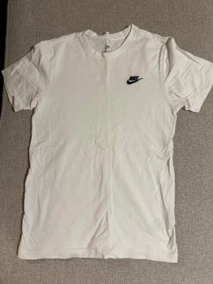 Vit Nike t-shirt med logga - Klassisk vit t-shirt från Nike med svart broderad logga på bröstet. Tillverkad i mjuk bomull och har en normal passform med korta ärmar och rund hals. Perfekt basplagg för en clean och sportig stil.
