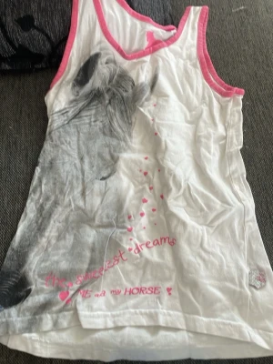 Vit linnetopp med hästmotiv och rosa kant - Säljer en vit linnetopp med stort hästmotiv i grått och texten 'the sweetest dreams' i rosa. Toppen har rosa kantband runt ärmhål och halsringning samt små rosa hjärtan som detalj. Perfekt för dig som älskar hästar!