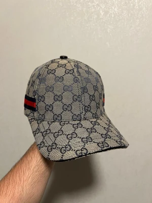 Gucci keps med GG-mönster - Snygg keps från Gucci i beige och mörkblått med det klassiska GG-monogrammet över hela kepsen. På sidan finns en röd och grön rand som signaturdetalj. Kepsen är tillverkad i bomull och har justerbar passform. Perfekt för dig som vill ha en exklusiv och trendig accessoar.