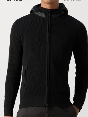 Svart stickad hoodie-cardigan - Snygg svart stickad cardigan med dragkedja och huva i quiltat material. Två fickor med dragkedja framtill och ribbade muddar vid ärmar och nederkant. Perfekt för lager på lager och en clean look.