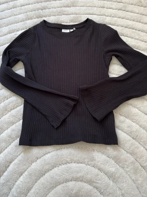 Svart ribbad långärmad topp - Svart långärmad topp från Monki med ribbad struktur. Modellen har rund halsringning och är tillverkad i ett mjukt, stretchigt material som sitter snyggt på kroppen. Perfekt att styla med jeans eller kjol.