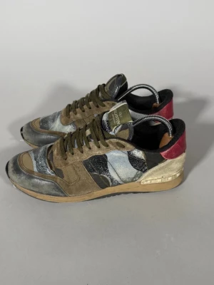Valentino kamouflage sneakers - Snygga sneakers från Valentino med kamouflagemönster i grönt, svart, beige och grått. Skorna har mockadetaljer, röda partier vid hälen och en robust gummisula. Klassisk låg modell med snörning och cool streetkänsla.
