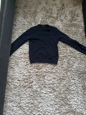 Mörkblå stickad tröja från Polo Ralph Lauren - Mörkblå stickad tröja från Polo Ralph Lauren med rund halsringning och diskret röd logga broderad på bröstet. Tröjan har långa ärmar och ribbade muddar vid ärmslut och nederkant. Perfekt för lager på lager och klassisk stil.