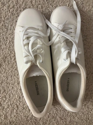 Vita sneakers från DinSko - Stilrena vita sneakers från DinSko med klassisk låg profil och vita snören. Skorna har en enkel och clean design i syntetmaterial som passar till det mesta. Perfekta för dig som gillar minimalistisk stil och vill ha ett par fräscha, vita sneakers i garderoben.