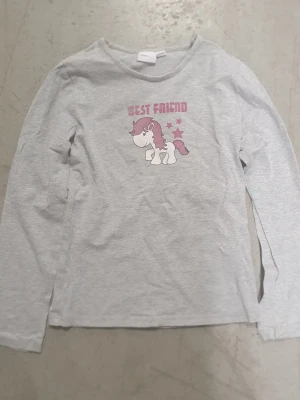 Tröja - Ljusgrå långärmad tröja med tryck av en söt enhörning och texten 'Best Friend' i rosa. Tröjan har rund halsringning och är gjord i mjukt bomullsmaterial. Perfekt för dig som gillar gulliga motiv och vill ha en bekväm vardagströja.