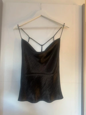 Svart satin linnetopp med tunna band - Snygg svart linnetopp i satin med tunna axelband och unik detalj i ryggen. Toppen har en lätt glansig finish och en lös passform som ger en trendig look. Perfekt att styla med jeans eller kjol för en cool vibe.