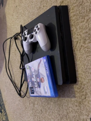 Vit DualShock 4 handkontroll PS4 - Playstation 4 Slim i svart med en vit DualShock 4-handkontroll. Konsolen har en stilren, platt design och är tillverkad i plast. Medföljer gör även kablar och spelet FIFA 23 i blå PS4-fodral. Perfekt för gaming och filmkvällar.