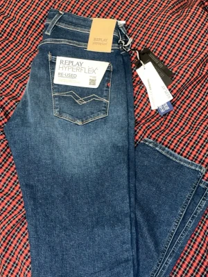Blå jeans från Replay Hyperflex - Snygga blå jeans från Replay Hyperflex med klassisk femficksdesign och vita kontrastsömmar. Jeansen har en rak passform och är tillverkade i stretchigt denimtyg för extra komfort. Perfekta för en avslappnad och trendig look. Det är storlek 27 men sitter som 28-29
