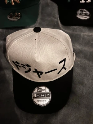 New Era 9FORTY keps med japansk text - Snygg 9FORTY keps från New Era med vit krona och svart skärm. Framtill finns broderad japansk text i svart. Justerbar passform med klassisk snapback-stil. Perfekt för dig som gillar streetwear och unika detaljer.