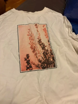 Vit t-shirt med blommigt tryck - Vit t-shirt med ett stort blommigt tryck i rosa och svart på framsidan. T-shirten har klassisk passform och rund hals. Perfekt för dig som gillar stilrena plagg med en twist.