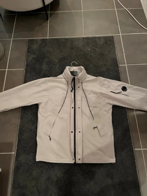 Vit vindjacka från C.P. Company - Snygg vit/grå vindjacka från C.P. Company med hög krage, svarta snören och dragkedja framtill. Jackan har en ikonisk linsdetalj på ärmen och praktiska fickor med dragkedja. Perfekt för dig som vill ha en stilren och funktionell jacka.