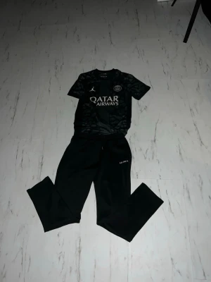 PSG x Jordan  - Säljer ett svart träningsset från PSG x Jordan med coolt mönster på tröjan och Qatar Airways-logga på bröstet. Setet består av en kortärmad tröja och matchande byxor med PSG-detaljer. Perfekt för fotbollsträning eller chill. Materialet är lätt och andas.