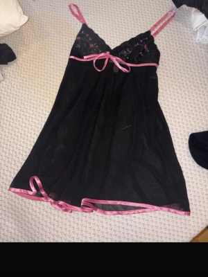 Svart nattlinne med spets och rosett - Sött svart nattlinne med tunna axelband och spets upptill. Dekorerat med rosa band, liten rosett under bysten och broderade blommor på spetsen. Nattlinnet har en lätt genomskinlig look och rosa kant nedtill. Perfekt för mysiga kvällar.