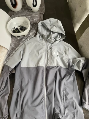 Under Armour jacka  - Snygg ljusgrå hoodie från Under Armour med dragkedja framtill och tvådelad design i olika grå nyanser. Hoodien har en huva med dragsko och praktiska fickor. Perfekt för en sportig och avslappnad stil. 