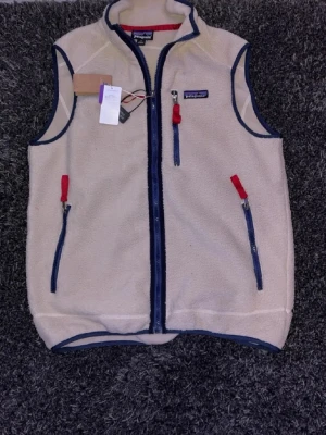 Beige fleeceväst från Patagonia - Snygg beige fleeceväst från Patagonia med marinblå detaljer och röda dragkedjor. Västen har hög krage, hel dragkedja framtill och tre fickor med dragkedja. Perfekt för lager-på-lager och outdoor-vibe, rekommenderas att vara runt 175-180 för den ska passa bäst! Priset kan diskuteras. Prislapp och påse följs med. 🤩 🙌 