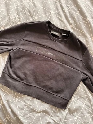 Svart cropped sweatshirt Calvin Klein - Snygg svart cropped sweatshirt från Calvin Klein med diskret logga broderad framtill. Tröjan har rund halsringning, långa ärmar och breda muddar. Perfekt till jeans eller kjol för en avslappnad look. Katt finns i hemmet. Liten i storleken.