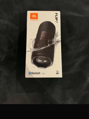 JBL Flip 7 trådlös högtalare svart - Säljer en JBL Flip 7 trådlös högtalare i svart. Högtalaren har en cylinderformad design med robust tygmaterial och gummidetaljer. Utrustad med Bluetooth, tydliga knappar för volym och uppspelning samt en praktisk bärrem. Vattentålig och har upp till 12 timmars batteritid. USB c 