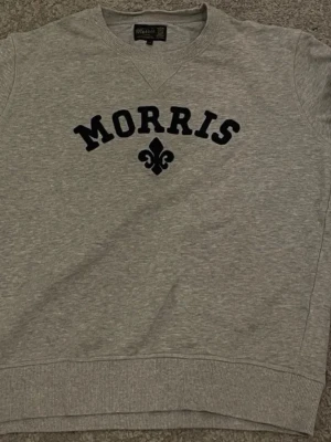 Grå sweatshirt från Morris - Snygg grå sweatshirt från Morris med svart text och dekorativt tryck på bröstet. Tröjan har rund halsringning, ribbade muddar och är tillverkad i mjukt bomullsmaterial. Perfekt för en avslappnad och stilren look.