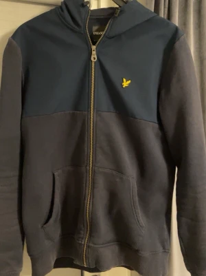 Mörkblå hoodie från Lyle & Scott - Snygg mörkblå hoodie från Lyle & Scott med dragkedja framtill och klassisk gul logga på bröstet. Hoodien har huva, känguruficka och ribbade muddar. Perfekt för en avslappnad och stilren look. Tillverkad i mjukt material för extra komfort.