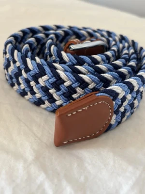 Blått flätat bälte med läderdetaljer 110 cm - Snyggt flätat bälte i blå, vit och marinblå färg med bruna läderdetaljer och silverfärgat spänne. Perfekt accessoar för att lyfta din outfit med en cool och avslappnad vibe.