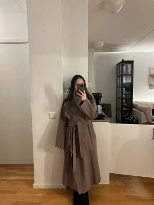 Brun lång ullkappa med knytskärp - Snygg och stilren brun kappa i lång modell med knytskärp i midjan. Kappan har breda slag och en rak passform som ger en trendig look. 100% ull! Nypris 5500kr