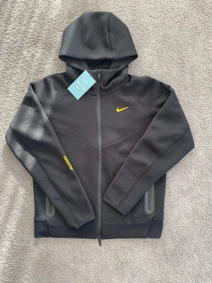 Svart Nike NOCTA hoodie med dragkedja - Svart hoodie från Nike NOCTA med gul logga på bröstet och detaljer på ärmen. Det står storlek M fast den passar bättre för storlek S