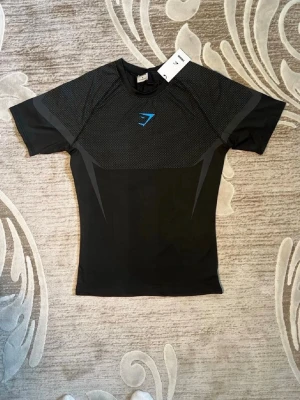 Svart träningströja från Gymshark - Snygg svart träningströja från Gymshark med korta ärmar och diskret mönstrad mesh upptill. Tröjan har en åtsittande passform och är tillverkad i ett stretchigt funktionsmaterial som andas. Gymshark-logga i blått på bröstet ger en sportig look.