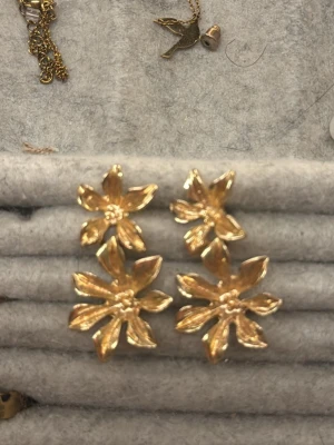 Guldiga blommiga statementörhängen - Säljer ett par stora örhängen i guldfärgad metall med tre blommor staplade på varandra. Örhängena har en glansig finish och en tydlig 3D-effekt som gör dem till riktiga statement pieces. Perfekta för dig som vill sticka ut med en unik accessoar.