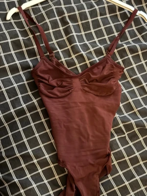 Seamless sculpt shapewear från Skims, storlek M - Säljer en vinröd shapewear bodysuit från Skims med smala, justerbara axelband och rynkade detaljer vid bysten. Tillverkad i ett mjukt och stretchigt material av polyamid och elastan. Perfekt för att skapa en snygg siluett under kläderna.