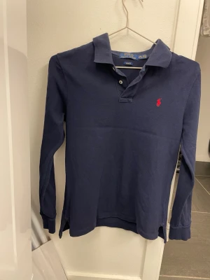 Marinblå långärmad skjorta Polo Ralph Lauren - Klassisk marinblå långärmad skjorta från Polo Ralph Lauren i slim fit-modell. Skjortan har krage, knappar framtill och den ikoniska röda logotypen broderad på bröstet. Tillverkad i mjuk bomull som känns skön mot huden.