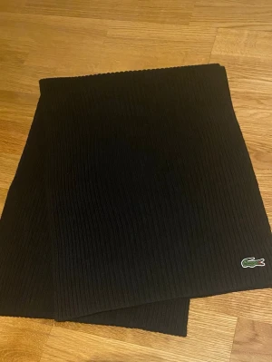 Svart ribbad halsduk från Lacoste - Snygg svart halsduk från Lacoste. 100% ull. Perfekt accessoar för att hålla värmen och lyfta din outfit. Ej använd!