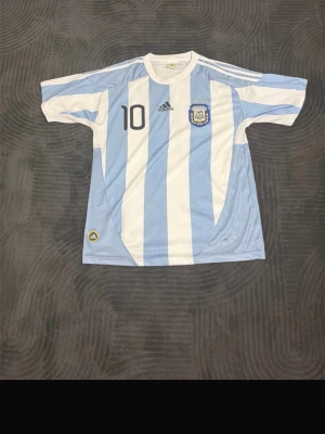 Argentina Messi tröja Adidas - Säljer en klassisk Argentina landslagströja från Adidas med ljusblå och vita ränder. Nummer 10 och Messi på ryggen, samt landslagets emblem på bröstet. Tröjan har korta ärmar och är tillverkad i lätt, ventilerande material.