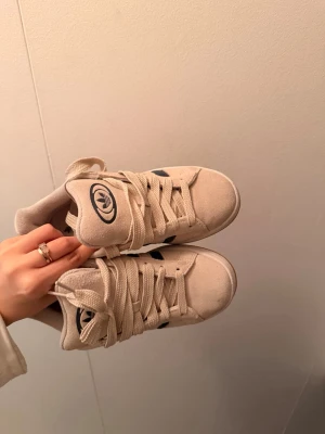 Adidas Campus - Snygga beige sneakers från Adidas med klassiska blå ränder på sidan och gummisula. Skorna har en låg profil, tjocka snören och Adidas-logga på plösen. Perfekta för en avslappnad streetstyle-look.