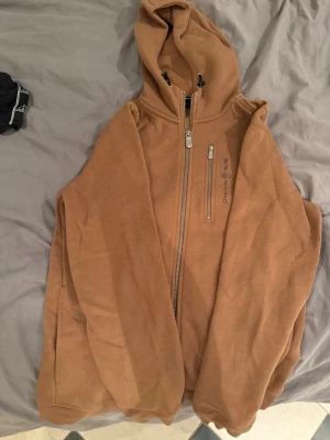 Brun hoodie med dragkedja från Sail Racing - Brun hoodie från Sail Racing med hel dragkedja och en extra bröstficka med zip. Hoodien har huva och är tillverkad i mjukt material, perfekt för chill dagar. Diskret logga på bröstet och klassisk passform.