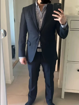 Kostym från Oscar Jacobson - Klassisk mörkgrå kostymset från Oscar Jacobson. Kavajen har två knappar framtill och fickor med lock. Stilren design med smal passform och diskret struktur i tyget. Perfekt för dig som vill ha en snygg och tidlös look. Både kavajen och byxorna är i utmärkt skick och har inga defekter. Kostymen är bara använd ett fåtal gånger. Skriv gärna för ytterligare frågor.