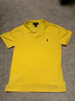 Gul pikétröja från Polo Ralph Lauren (14-16) - Klassisk gul pikétröja från Polo Ralph Lauren med blå broderad logga på bröstet. Tröjan har korta ärmar, krage och två knappar vid halsen. Tillverkad i bomull och har en custom fit passform.
