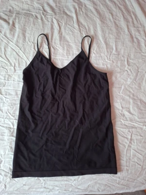 Svart linnetopp med smala axelband - En enkel och stilren svart linnetopp med smala axelband och v-ringad hals. Perfekt att matcha med jeans eller kjol för en clean look. Tillverkad i mjukt material som känns skönt mot huden.