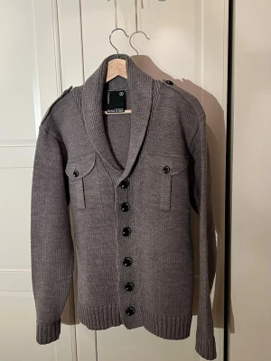 Grå cardigan med knappar Soulstar - Stilren grå stickad cardigan från Soulstar med stora svarta knappar, två bröstfickor med lock och axelklaffar. Koftan har ribbade muddar och en bred sjalkrage som ger en avslappnad men snygg look. Perfekt till för lager-på-lager. Storlek S men passar M. Inga defekter. Skriv vid minsta fundering👋
