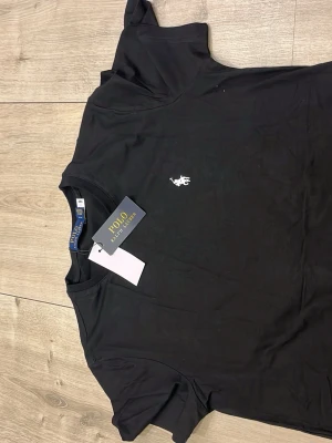 Svart t-shirt från Polo Ralph Lauren - Snygg svart t-shirt från Polo Ralph Lauren med klassisk vit logga broderad på bröstet. T-shirten har rund halsringning och korta ärmar. Tillverkad i mjuk bomull som känns skön mot huden. Perfekt basplagg till garderoben.