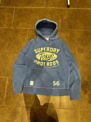 Blå Superdry hoodie - Säljer en blå hoodie från Superdry med stort gult Tokyo Hot Rods-tryck på bröstet och siffran 56 nedtill. Klassisk känguruficka, ribbade muddar och dragsko i huvan. Perfekt för dig som gillar streetwear och vill ha en skön och snygg hoodie.