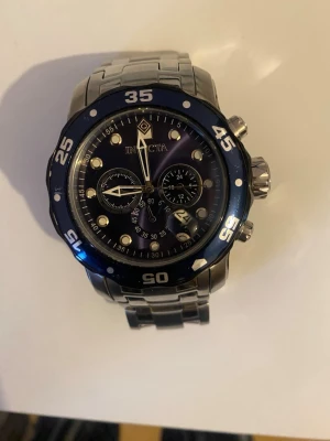 Invicta silverfärgad kronografklocka - Stilren kronografklocka från Invicta med robust silverfärgad länkarmband i metall. Urtavlan är svart med blå ram och har flera subdials samt tydliga siffror och markeringar. Perfekt för dig som gillar sportig och modern design.