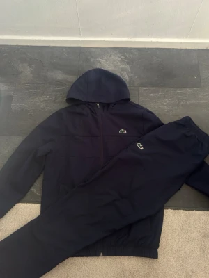 Lacoste trackie - Snyggt marinblått loungewear-set från Lacoste Sport med huva och dragkedja på tröjan samt matchande byxor. Setet har den klassiska Lacoste-loggan på både tröja och byxa. Regular fit och tillverkat i mjukt material, perfekt för chill dagar hemma.