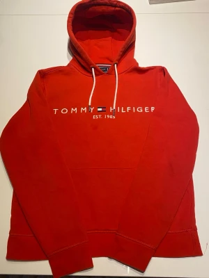 Röd hoodie Tommy Hilfiger - Snygg röd hoodie från Tommy Hilfiger med vit logga och flagga broderad på bröstet. Tillverkad i mjuk ekologisk bomull och har klassisk huva med vita snören. Perfekt för en avslappnad och trendig stil.