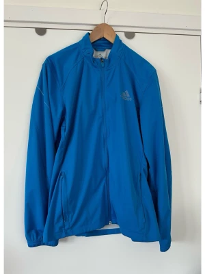Blå träningsjacka från Adidas, storlek M - Blå Adidas Climaproof träningsjacka med dragkedja framtill och hög krage. Jackan har två fickor med dragkedja i sidorna och en Adidas-logotyp på bröstet. Ärmslut och nederkant är elastiska. Praktiska vädrings-skenor bak på ryggen. Reflex-märkningen på ärmarna har gått bort, se bild, men inget som syns eller som man tänker på. Annars i toppenskick, som ny!