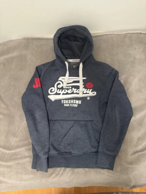 Mörkblå Superdry hoodie med tryck - Snygg blå hoodie från Superdry med vit logga och texten 'Yokohama High Flyers' på bröstet. Har stor huva med vita snören och en klassisk känguruficka framtill. Röd detalj på ärmen och mjukt material som känns skönt mot huden. PRIS KAN DUSKITERAS