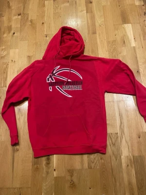 Röd hoodie Eastside Lutheran Basketball stl M  - Röd hoodie med tryck av en basketboll och texten 'Eastside Lutheran Basketball' på bröstet. Klassisk känguruficka framtill och justerbar huva med dragsko. Perfekt för dig som gillar sportig stil och basketvibbar.
