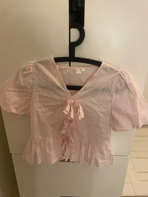 Ljusrosa blus med volang och knyt - Supersöt ljusrosa blus med v-ringning, puffärmar och volangkant nedtill i storlek 146/152. Framtill finns dekorativa knytband som ger en romantisk vibe. Perfekt till jeans eller kjol för en trendig look.