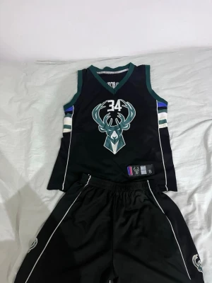 Milwaukee Bucks basketset svart - Snyggt basketset med Milwaukee Bucks tryck, nummer 34 och stor logga på bröstet. Svart linne med gröna och vita detaljer samt matchande shorts. Perfekt för basketträning eller streetstyle. Materialet är lätt och andas, vilket gör det bekvämt att spela i. Namn på ryggen är Giannis Antetokounmpo om ni vill se bild bara skicka ett meddelande 